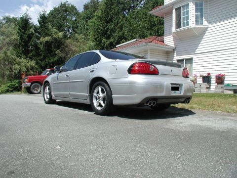 2002 Pontiac Grand Prix - The rear passenger side of a silver sedan is shown parked on an asphalt surface in front of a white house. A red pickup truck is visible in…