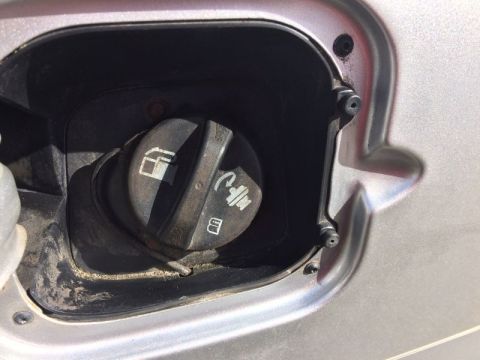 2002 Pontiac Grand Prix - Close-up of a silver car's fuel filler door, showing the black gas cap with fuel and emissions symbols. The surrounding area is dirty.