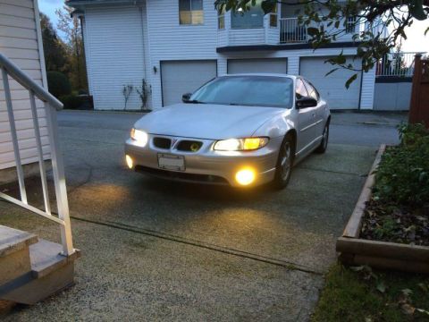2002 Pontiac Grand Prix - A silver Pontiac Grand Prix is parked in a driveway in front of a white house with multiple garage doors. The car's headlights and fog ligh…