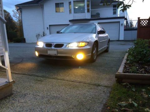 2002 Pontiac Grand Prix - A silver Pontiac Grand Prix sedan is parked in a driveway at dusk, with its headlights and fog lights illuminated. The car is positioned in…