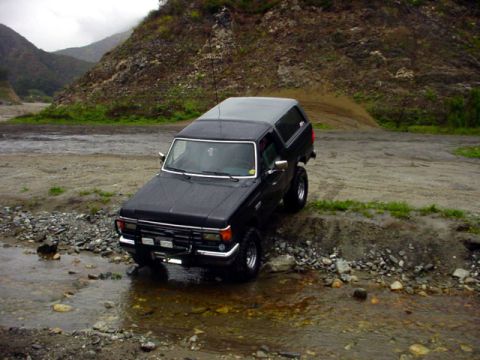 1988 Ford Bronco - Off-road SUV navigates rocky terrain and shallow water on a cloudy day.