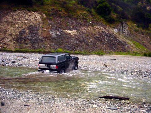 1988 Ford Bronco - A Ford Bronco navigates a shallow, rocky river with a steep, vegetated hillside in the background.