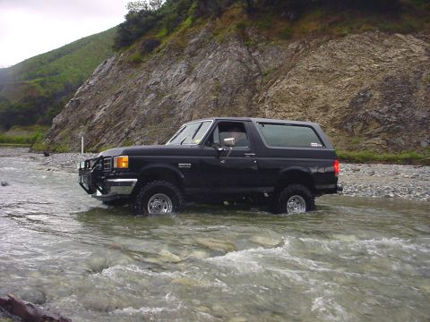1988 Ford Bronco - A black 1987 Ford Bronco navigates a shallow river with a rocky bottom and a steep, vegetated hillside in the background.