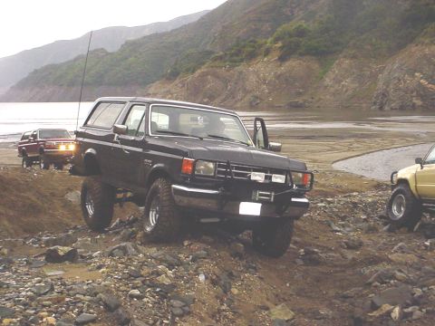 1988 Ford Bronco - A black Ford Bronco navigates a rocky, off-road terrain near a body of water, with other vehicles visible in the background.