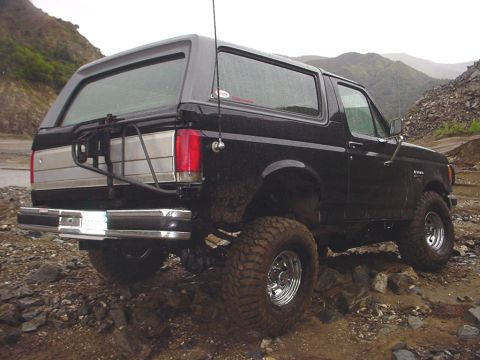 1988 Ford Bronco - This black Ford Bronco is shown from the rear on a muddy, rocky terrain with hills in the background.