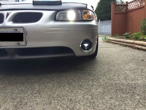2002 Pontiac Grand Prix - The front bumper and grille of a silver Pontiac Grand Prix are shown, with the headlights and a fog light illuminated. The vehicle is parke…