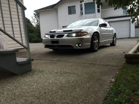 2002 Pontiac Grand Prix - A silver Pontiac Grand Prix is parked in a driveway in front of a white house. The car has a black bra on the front bumper and its headligh…