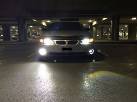 2002 Pontiac Grand Prix - The front of a silver sedan with its headlights and fog lights on is parked in a dimly lit parking garage. The wet asphalt reflects the bri…