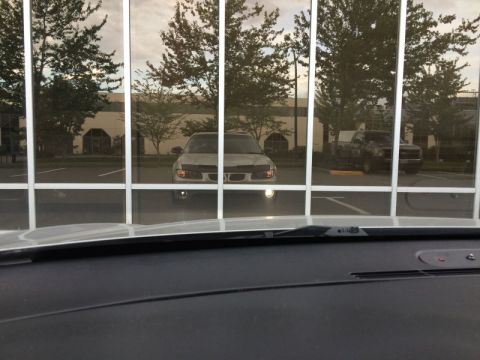 2002 Pontiac Grand Prix - The front of a silver sedan is reflected in a building's windows. The car is parked in a lot with trees and another vehicle visible in the…