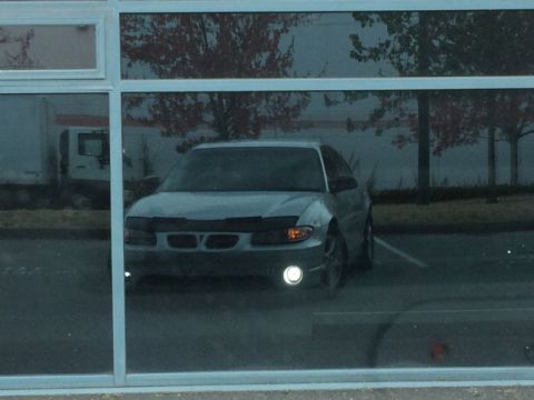 2002 Pontiac Grand Prix - The front of a silver sedan is visible through a reflective window, showing its headlights and grille. The car is parked in an outdoor lot…