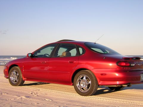 1997 Ford Taurus - A red 1997 Ford Taurus is seen from a three-quarter front view on a beach with the ocean and sky behind it.
