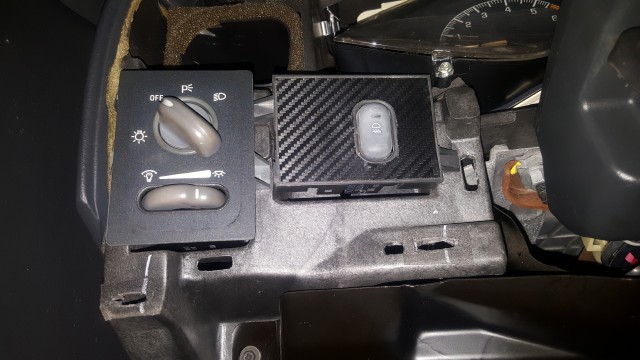 Vehicle dashboard showing headlight control knob and a button with carbon fiber trim.