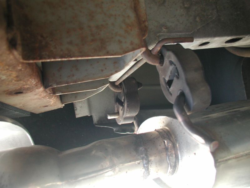 Underside of a vehicle showing rusted frame and exhaust hangers with rubber isolators.
