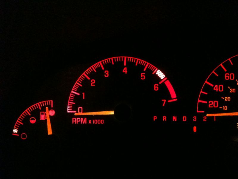Car dashboard at night with red illuminated tachometer, fuel gauge, and speedometer.