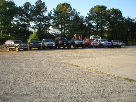 1989 Ford Bronco - A collection of SUVs and trucks are parked in a gravel lot under a clear sky.