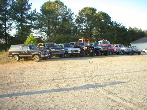 1989 Ford Bronco - Several SUVs and trucks are parked in a line on a gravel lot with trees in the background.