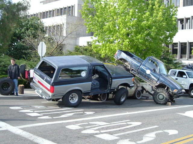 Gray Ford Bronco with rear lifted and a blue pickup tipped nose-down against it on a city street