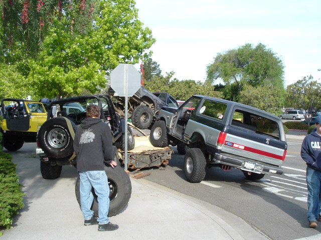 Lifted two-tone Ford Bronco climbing a wooden ramp obstacle at a parking-lot 4x4 event with onlookers