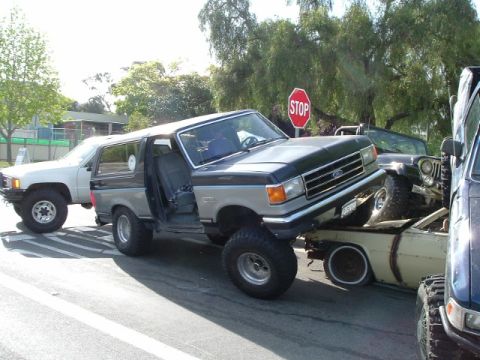 1989 Ford Bronco - A lifted Ford Bronco is precariously perched on top of a sedan, with other vehicles around.