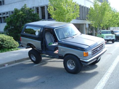 1989 Ford Bronco - A lifted, two-tone Ford Bronco is parked on the street with its driver's door open, while other vehicles are visible in the background.