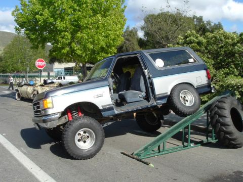 1989 Ford Bronco - This lifted 1989 Ford Bronco is shown with its doors open, ascending a ramp with a large tire nearby.