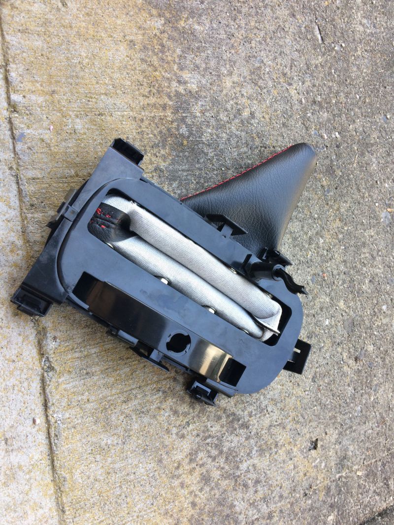 Car part, likely airbag module, with black plastic, folded fabric, and red stitching on leather trim, on concrete.