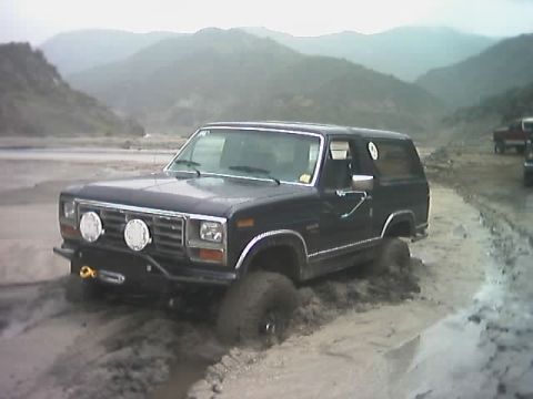 1985 Ford Bronco - A dark blue Ford Bronco is stuck in mud with mountains in the background, and another truck is visible.