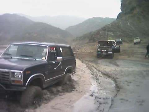 1985 Ford Bronco - A dark blue SUV is stuck in the mud on a rough, mountainous road with several other vehicles in the background.