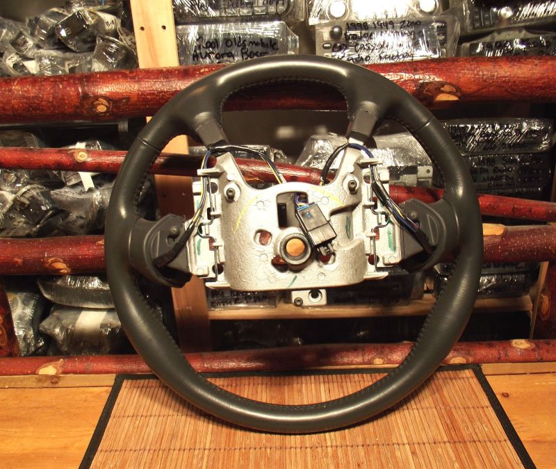 Black leather steering wheel with silver hub, mounted on a wooden rack with car parts.