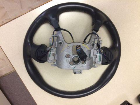 2002 Pontiac Grand Prix - A black leather steering wheel with a silver metal hub and wiring is shown from a top-down perspective on a light-colored surface.