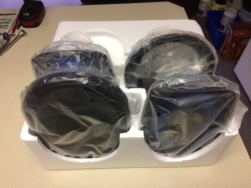 Four black car speakers in plastic wrap and styrofoam packaging.