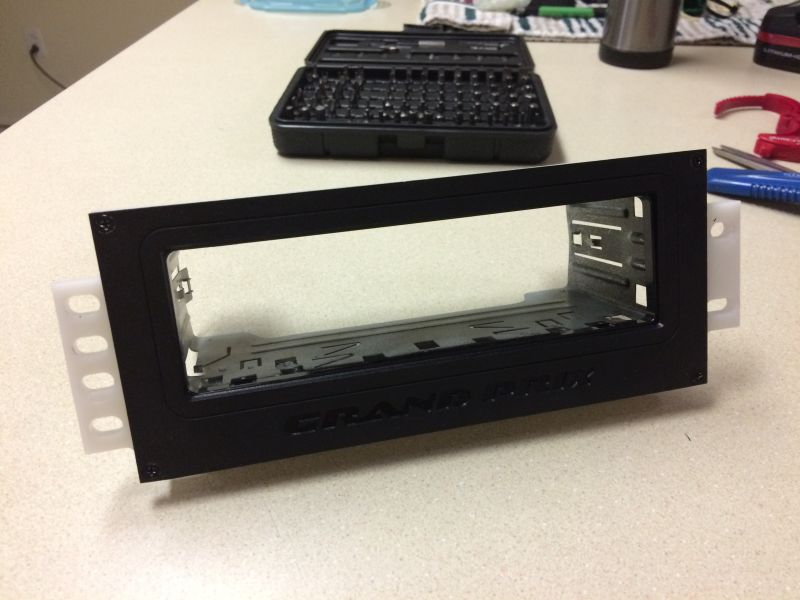 Black 1-DIN car stereo mounting bracket with white side wings, text "GRAND AM" visible.
