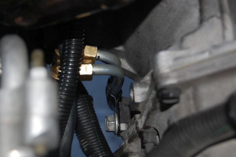 Close-up of two metal fuel lines with brass fittings connected to a black corrugated hose on an engine.