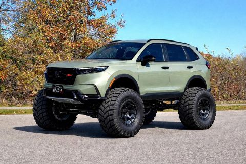 2026 Honda CR-V - A lifted, light green SUV with oversized off-road tires is parked on an asphalt road with trees and a clear blue sky in the background.