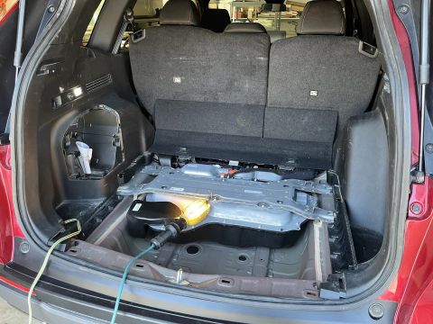 2026 Honda CR-V - The interior of a red SUV's cargo area is shown with the floor panel removed, revealing the battery pack and wiring. A yellow work light is…