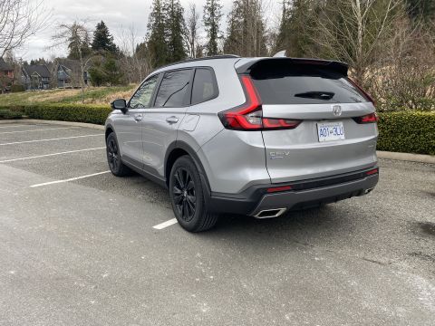 2026 Honda CR-V - The rear driver's side of a silver Honda CR-V is parked in an empty parking lot with trees and houses in the background.