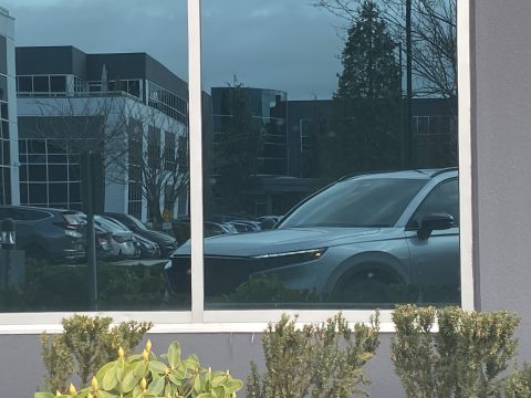 2026 Honda CR-V - The reflection in a building's window shows the front 3/4 view of a light silver SUV parked in a lot. The vehicle has a sleek design with s…