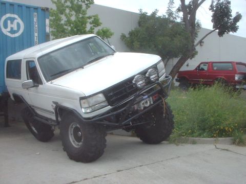 1995 Ford Bronco - A modified white Ford Bronco is parked next to a blue shipping container, with a red Bronco visible in the background.