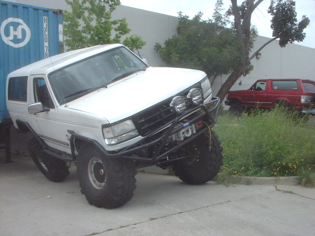 White lifted Ford Bronco flexing front suspension on a curb, black tube bumper with round lights.