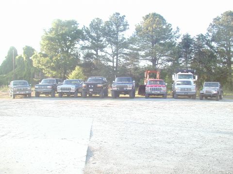 1988 Ford Bronco - Several trucks and SUVs are parked in a line outdoors under a bright sky.