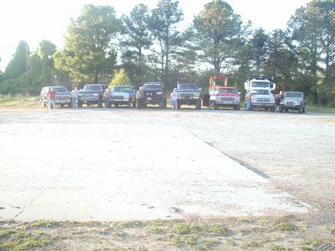 1988 Ford Bronco - A collection of SUVs and trucks are lined up in a parking lot with trees in the background.