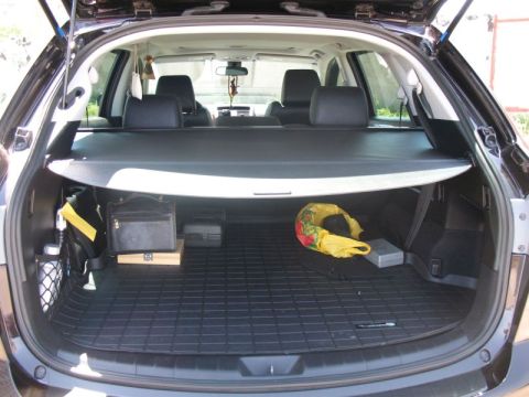 2009 Mazda CX-9 - The open rear cargo area of a dark colored SUV is shown, featuring a retractable cargo cover and various items stored on the floor mat. Vis…