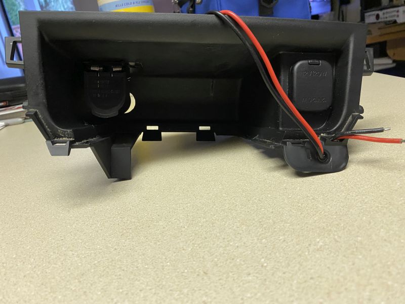 Black plastic automotive dash panel with two power outlets and wires.