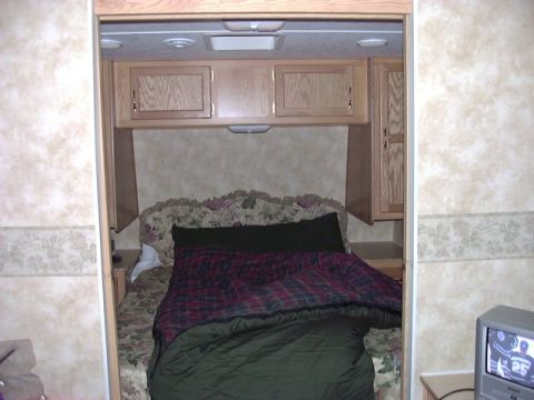 1992 Ford Bronco - Interior view of an RV bedroom a bed with a plaid blanket and sleeping bag, surrounded by wood cabinetry.