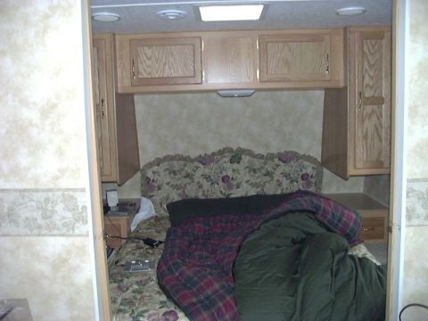 1992 Ford Bronco - The interior of an RV bedroom shows a made bed with a floral headboard and wooden cabinets overhead.