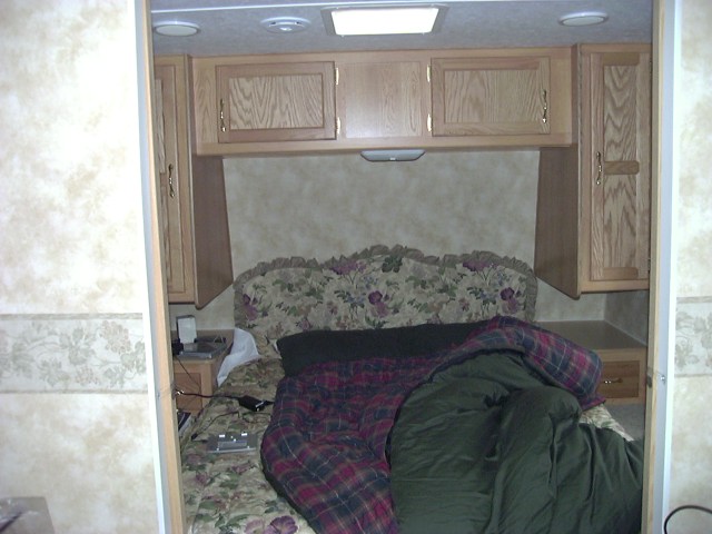 RV bedroom with a floral headboard, plaid comforter, and wooden cabinets above the bed