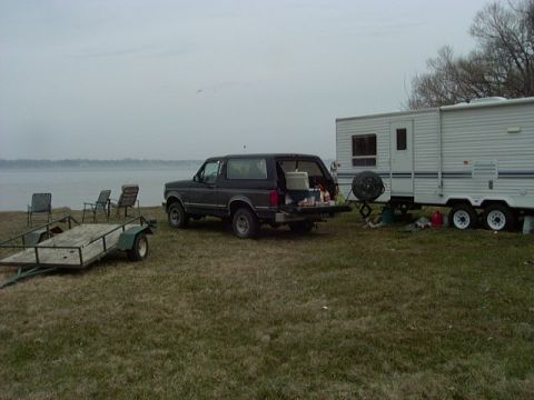 1992 Ford Bronco - A 1992 Ford Bronco is parked near a trailer and RV with the tailgate open, suggesting a campsite setup by the water.