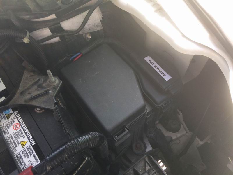 Vehicle engine bay with fuse box labeled DRL BYPASS and car battery.