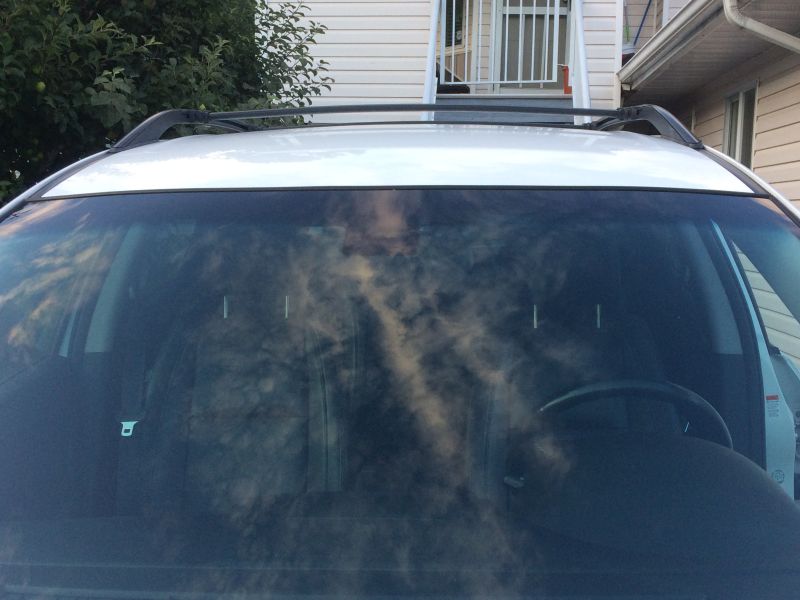 White SUV windshield and roof rack with sky reflection, parked in front of a house.