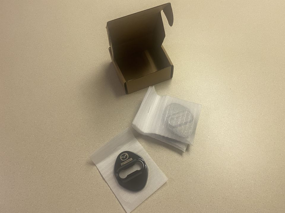 Two black Mazda door lock covers on a countertop, one in a bag.